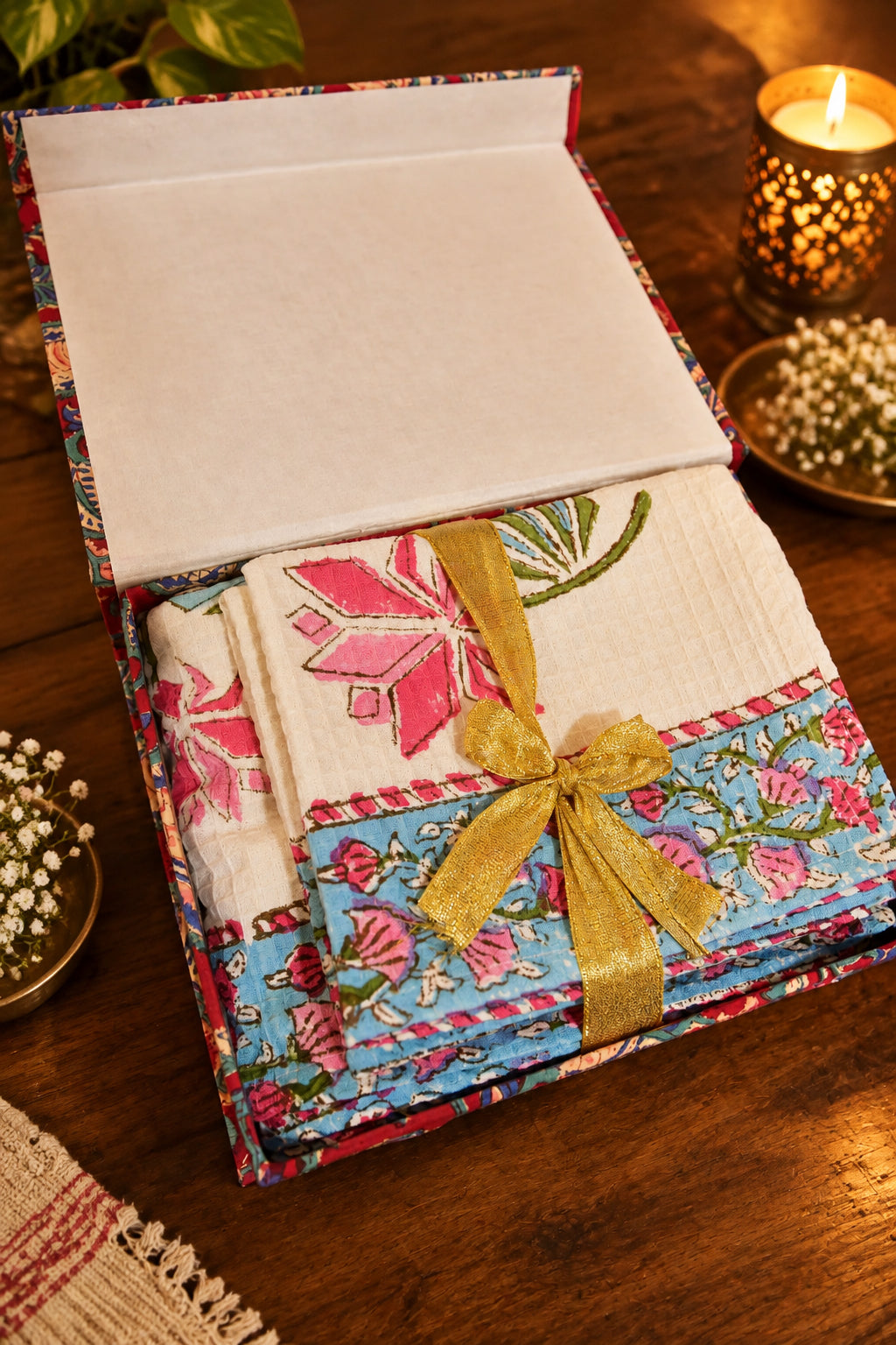A folded textile gift set in a patterned box with a white interior, tied with a gold ribbon. The fabric features a white textured section with pink floral embroidery and a blue floral border.
