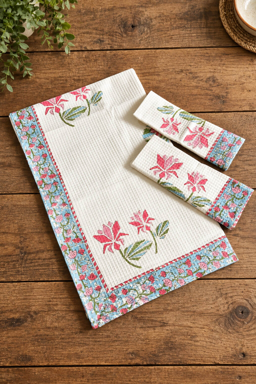 A matching kitchen towel set with a white waffle-weave texture, featuring pink floral motifs and a blue floral border, displayed on a rustic wooden table.