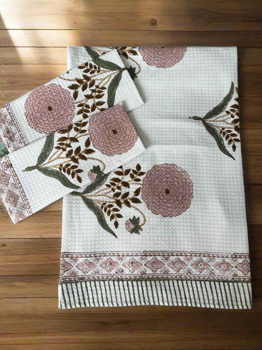 A white textured cotton tablecloth with a floral block print pattern in dusty pink and brown, laid flat on a wooden floor alongside matching napkins.