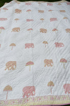A white quilted blanket featuring a repeating pattern of pink elephants and pink-and-beige umbrellas, finished with a decorative floral border.