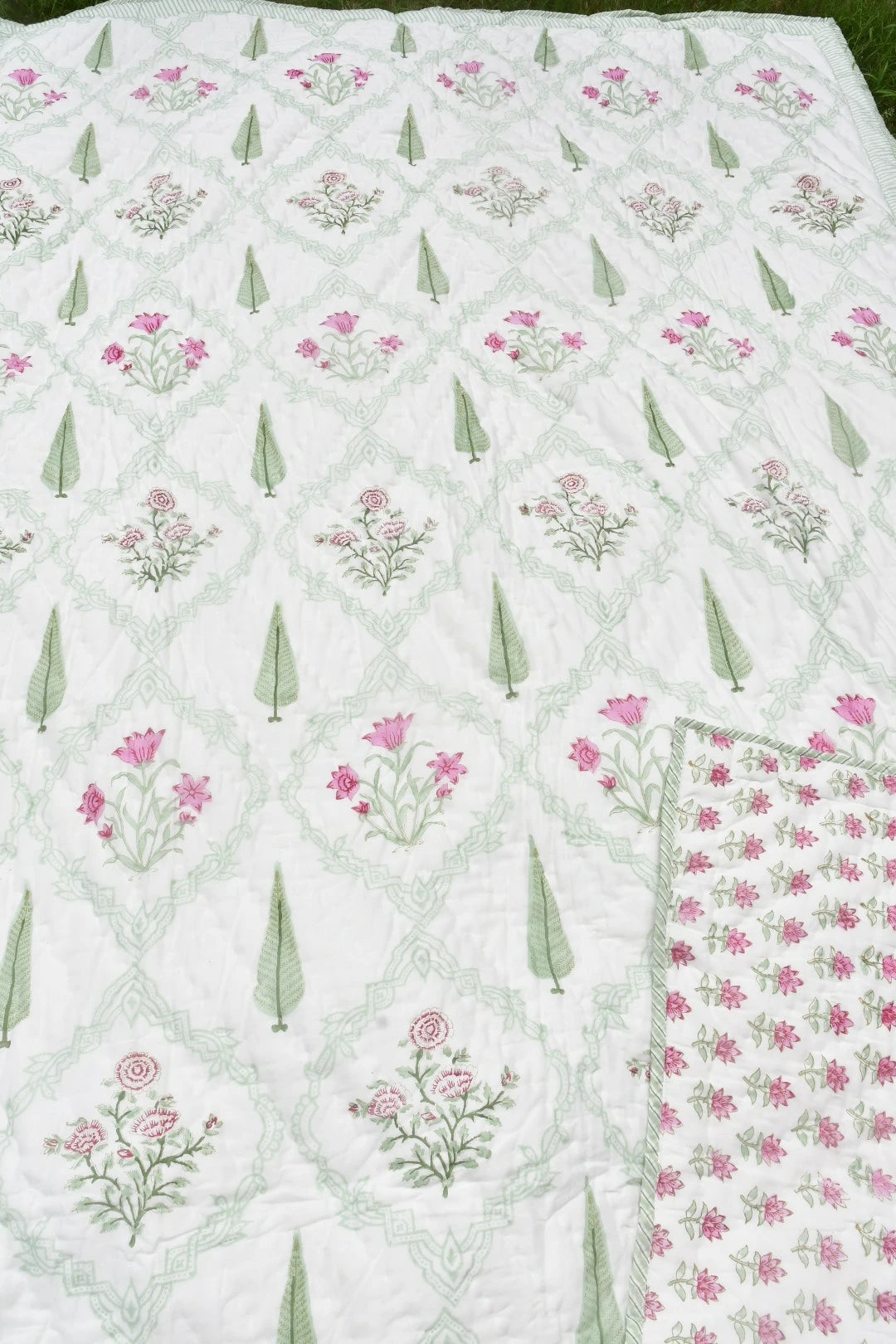 A white quilted blanket with a repeating pattern of pink flowers, green leaves, and a light green trellis design, shown with a folded corner revealing the reverse side.