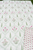 A white quilted blanket with a repeating pattern of pink flowers, green leaves, and a light green trellis design, shown with a folded corner revealing the reverse side.