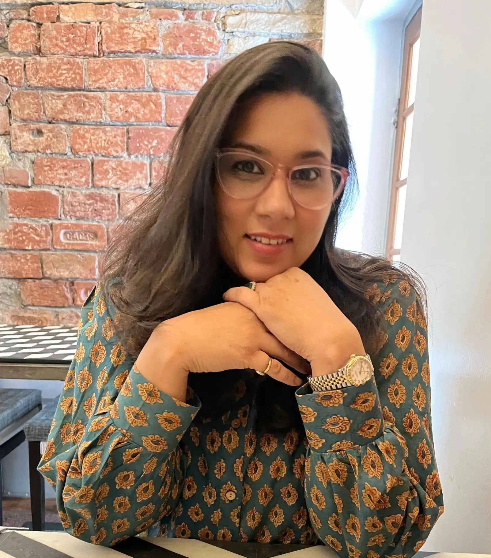 Woman wearing glasses and a patterned blouse sitting against a brick wall.