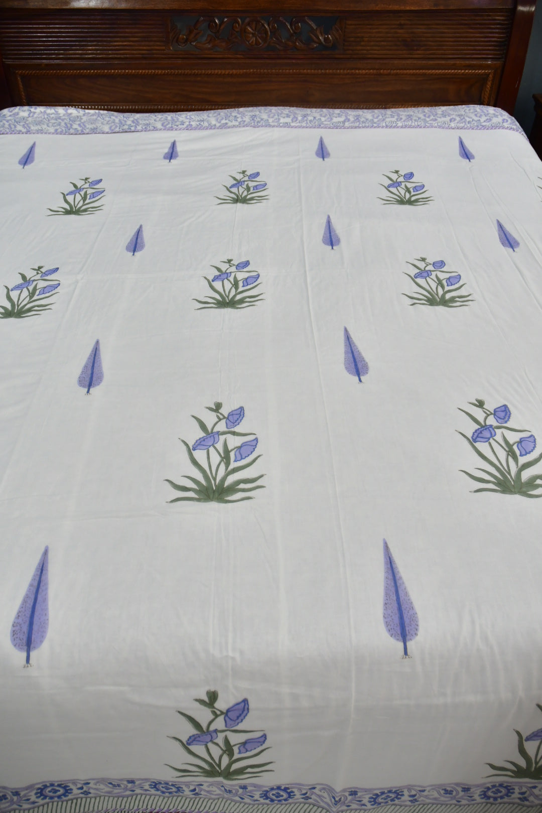 A white cotton sheet or bedspread featuring a repeating pattern of blue-purple flowers with green stems and blue teardrop motifs, with a blue patterned border at the top and bottom.