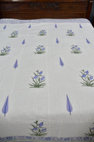 A white cotton sheet or bedspread featuring a repeating pattern of blue-purple flowers with green stems and blue teardrop motifs, with a blue patterned border at the top and bottom.