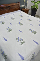 A white bedspread or comforter with a repeating pattern of blue irises and purple leaves, styled on a bed with a dark wood headboard in a bedroom setting.