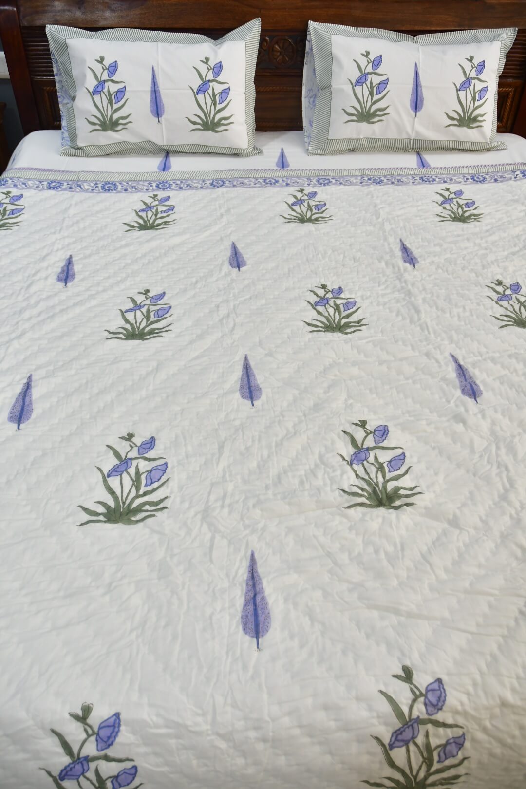 An off-white quilted bedsheet with embroidered blue and green floral motifs and a blue border, shown on a bed with two matching pillowcases.