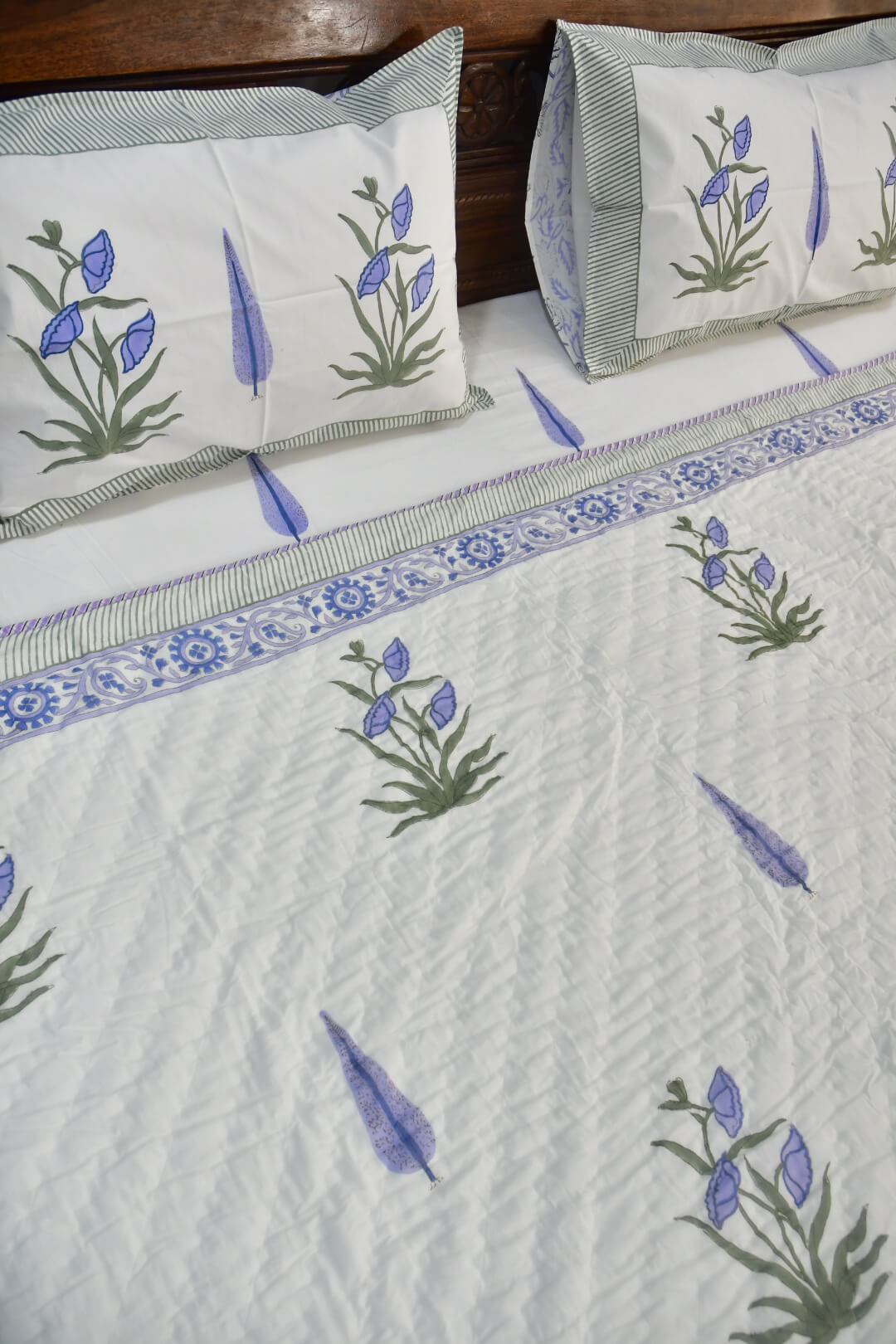 A close-up of a quilted white bedding set featuring a blue and green floral pattern, matching pillowcases, and a decorative border, styled on a bed with a dark wooden headboard.