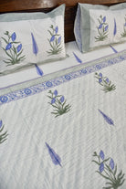 A close-up of a quilted white bedding set featuring a blue and green floral pattern, matching pillowcases, and a decorative border, styled on a bed with a dark wooden headboard.