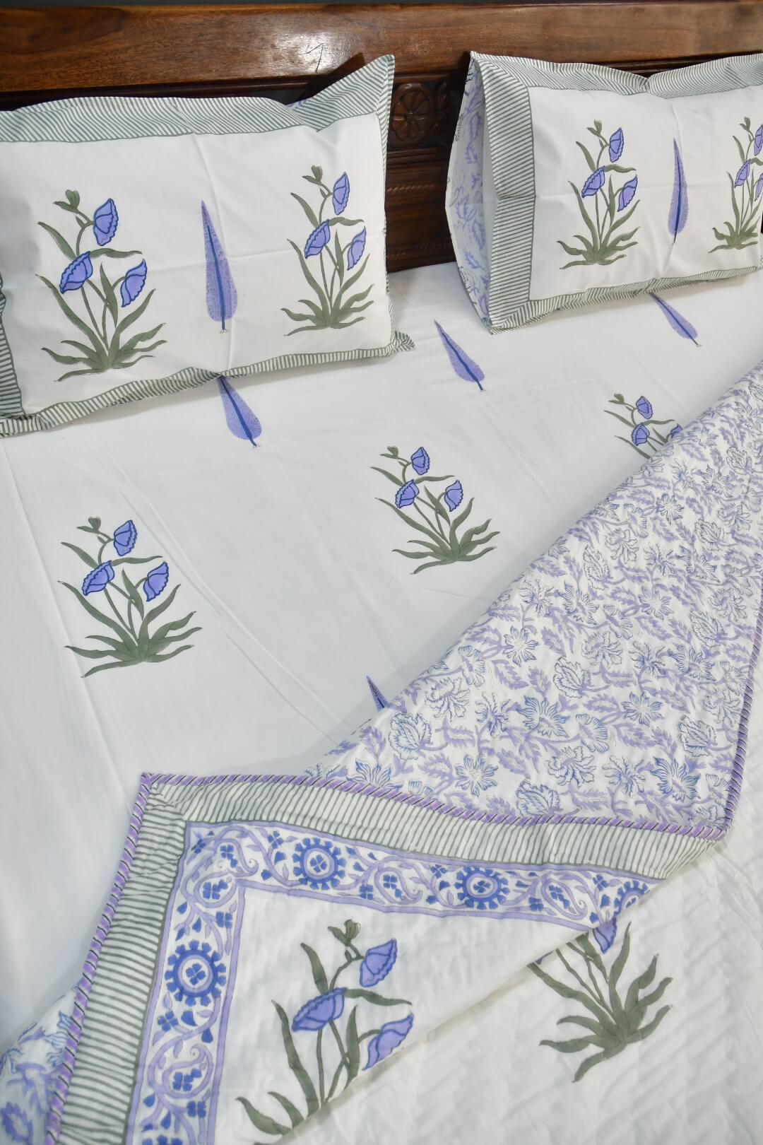 A white cotton bedding set featuring block-printed blue lotus flowers and green leaves, with striped and floral patterned borders in blue, green, and white.