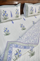A white cotton bedding set featuring block-printed blue lotus flowers and green leaves, with striped and floral patterned borders in blue, green, and white.