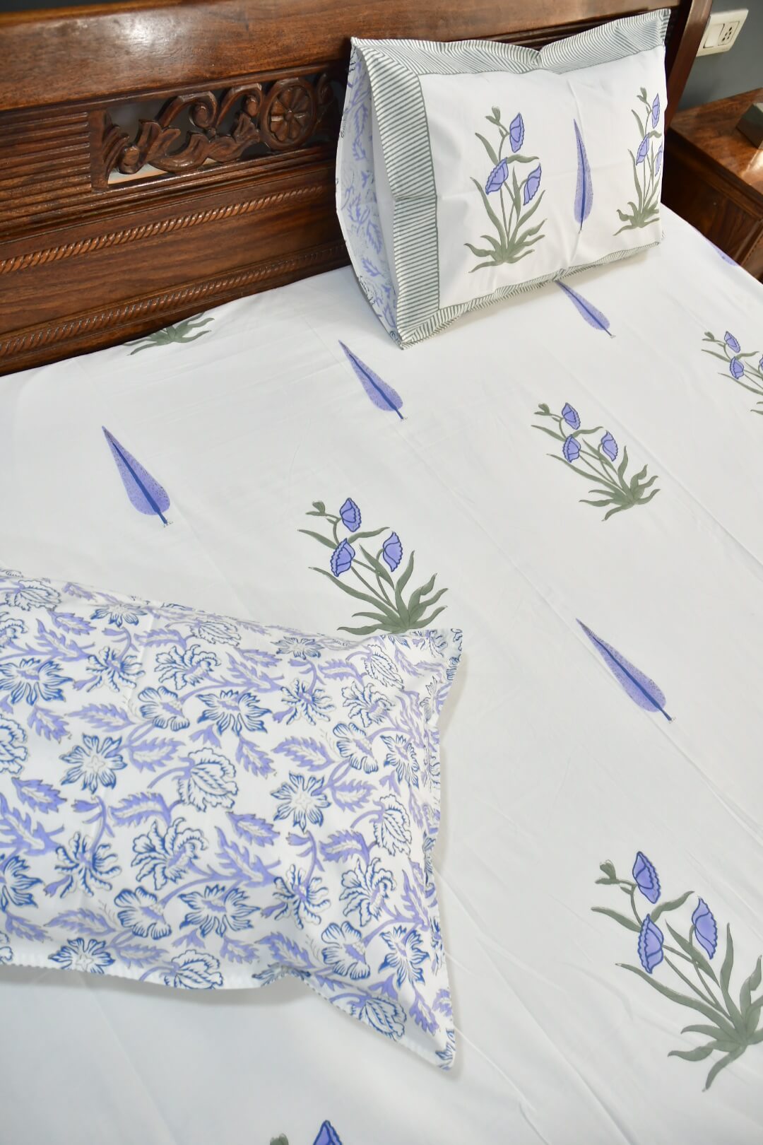 A close-up of a bed made with a white bedsheet featuring blue and green floral and leaf patterns, including a patterned pillowcase and a matching pillow on a wooden bed frame.