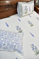 A close-up of a bed made with a white bedsheet featuring blue and green floral and leaf patterns, including a patterned pillowcase and a matching pillow on a wooden bed frame.