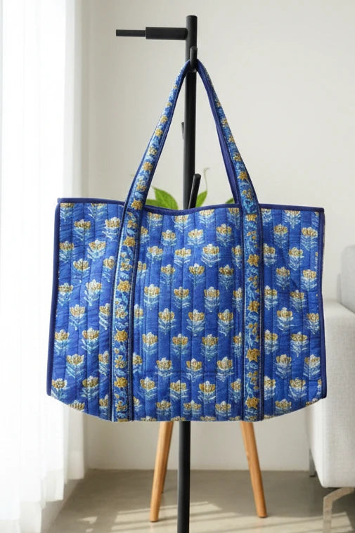 A blue quilted tote bag with a yellow and white floral pattern, hanging from a black coat rack in a bright room.