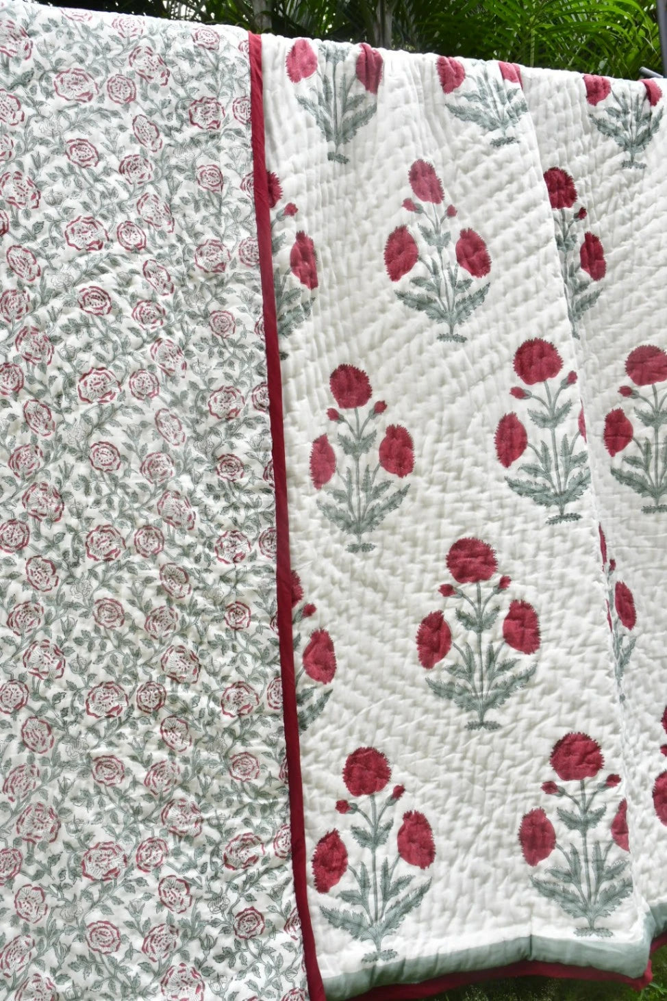 Two quilted fabric panels with floral patterns hanging side by side outdoors. One panel has a dense small floral print in muted red and gray-green on a light background, and the other has larger red floral motifs with gray-green stems on a white background, both with red edging.