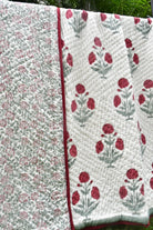 Two quilted fabric panels with floral patterns hanging side by side outdoors. One panel has a dense small floral print in muted red and gray-green on a light background, and the other has larger red floral motifs with gray-green stems on a white background, both with red edging.