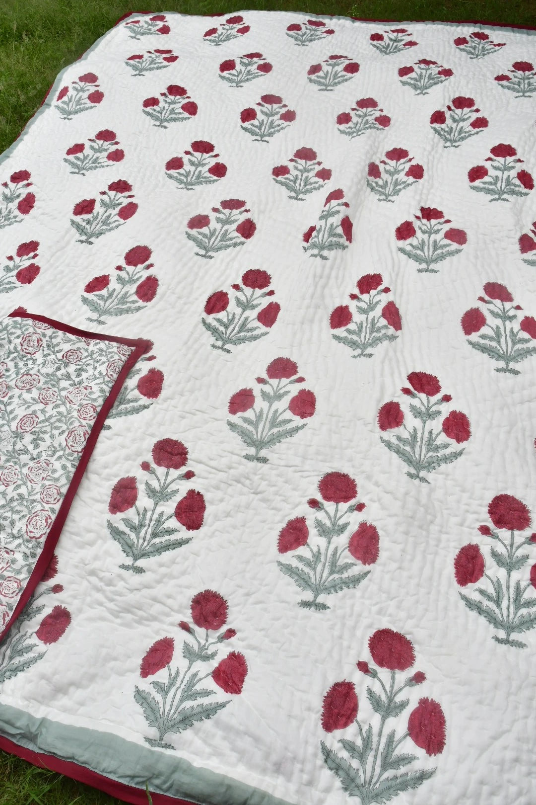 A white quilted blanket with a repeating red and gray floral block print pattern, laid out on green grass. A folded corner of the blanket reveals a red border and a matching patterned reverse side.
