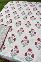 A white quilted blanket with a repeating red and gray floral block print pattern, laid out on green grass. A folded corner of the blanket reveals a red border and a matching patterned reverse side.