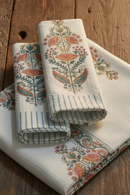 Three folded cotton kitchen towels with blue stripes and orange, teal, and green floral block prints, arranged on a rustic wooden table.
