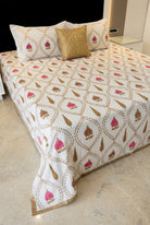 A bed dressed in a cream-colored cotton sheet with brown and pink block-printed motifs, styled with decorative pillows in a bedroom setting. The brand name ALBELI is visible as an overlay in the bottom-left corner.