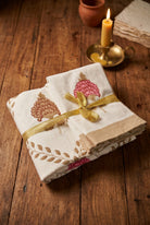 A folded white fabric item, likely a napkin or small cloth, decorated with embroidered floral motifs in pink and brown, tied with a shiny gold ribbon, resting on a dark wooden surface.