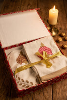 A folded gift set of white embroidered cotton textiles, likely handkerchiefs or napkins, presented in an open decorative box with a red patterned lining and tied with a gold ribbon. The textiles feature pink and gold floral embroidery and gold leaf motifs.