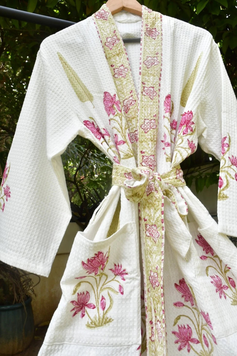 A white waffle-knit robe with pink floral embroidery and a beige patterned border, tied at the waist with a matching tie belt, hanging outdoors against a green foliage background.