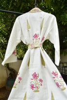 An off-white cotton kimono robe with pink floral embroidery and gold leaf patterns, hanging on a black clothesline outdoors with green foliage in the background.