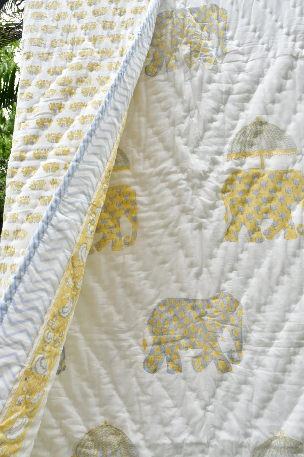A close-up of a white quilted blanket featuring yellow elephant motifs with patterned umbrellas, a blue and white chevron border, and a yellow patterned reverse side.