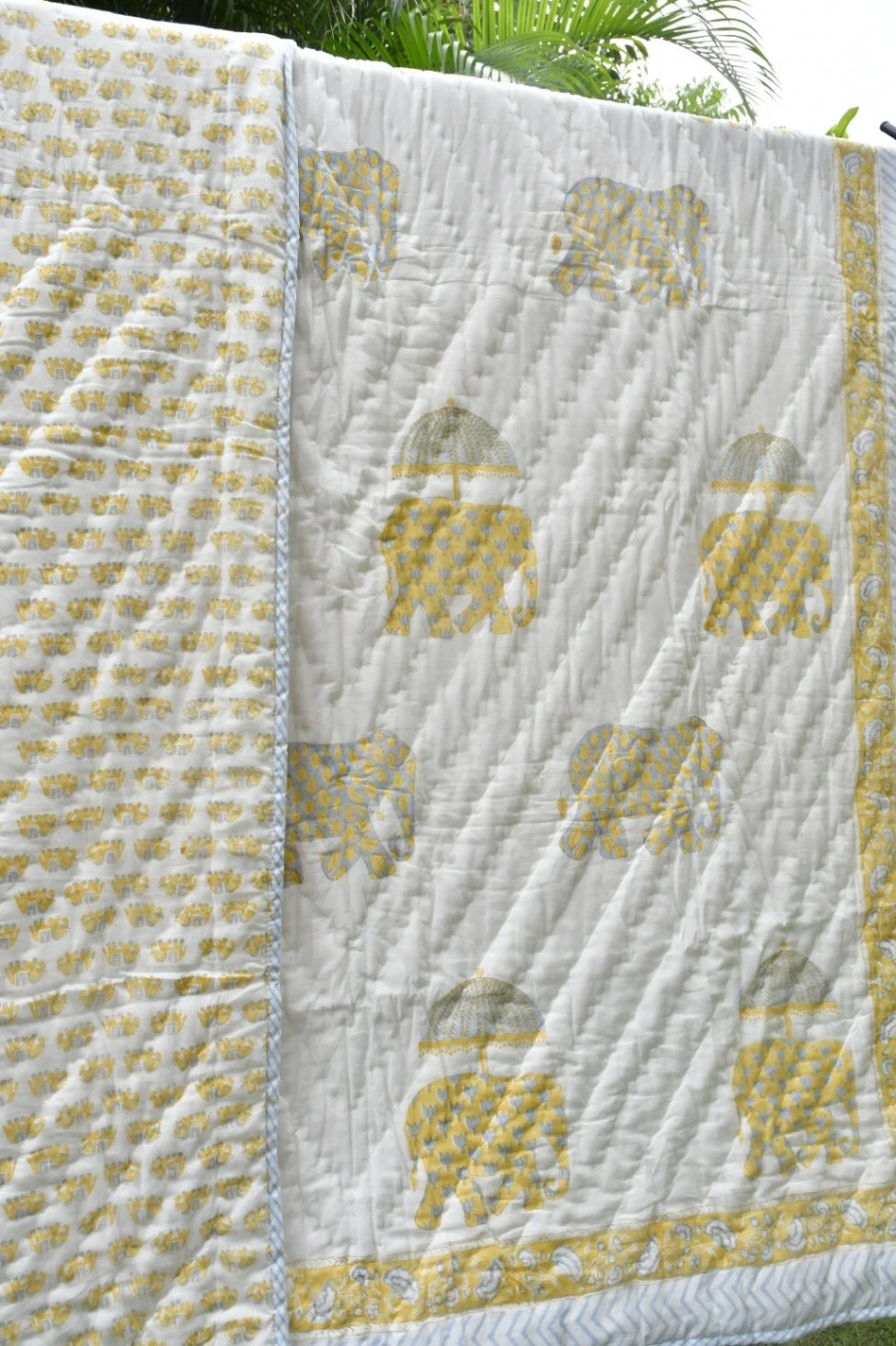 A white quilted blanket featuring a repeating pattern of gold elephants and a matching gold border, shown outdoors with green foliage visible in the background.