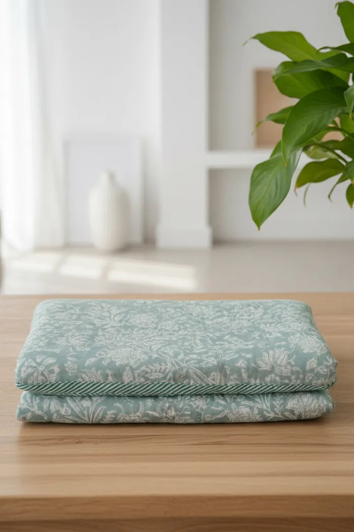 Folded teal blanket with floral pattern on a wooden surface, plant and white wall in the background