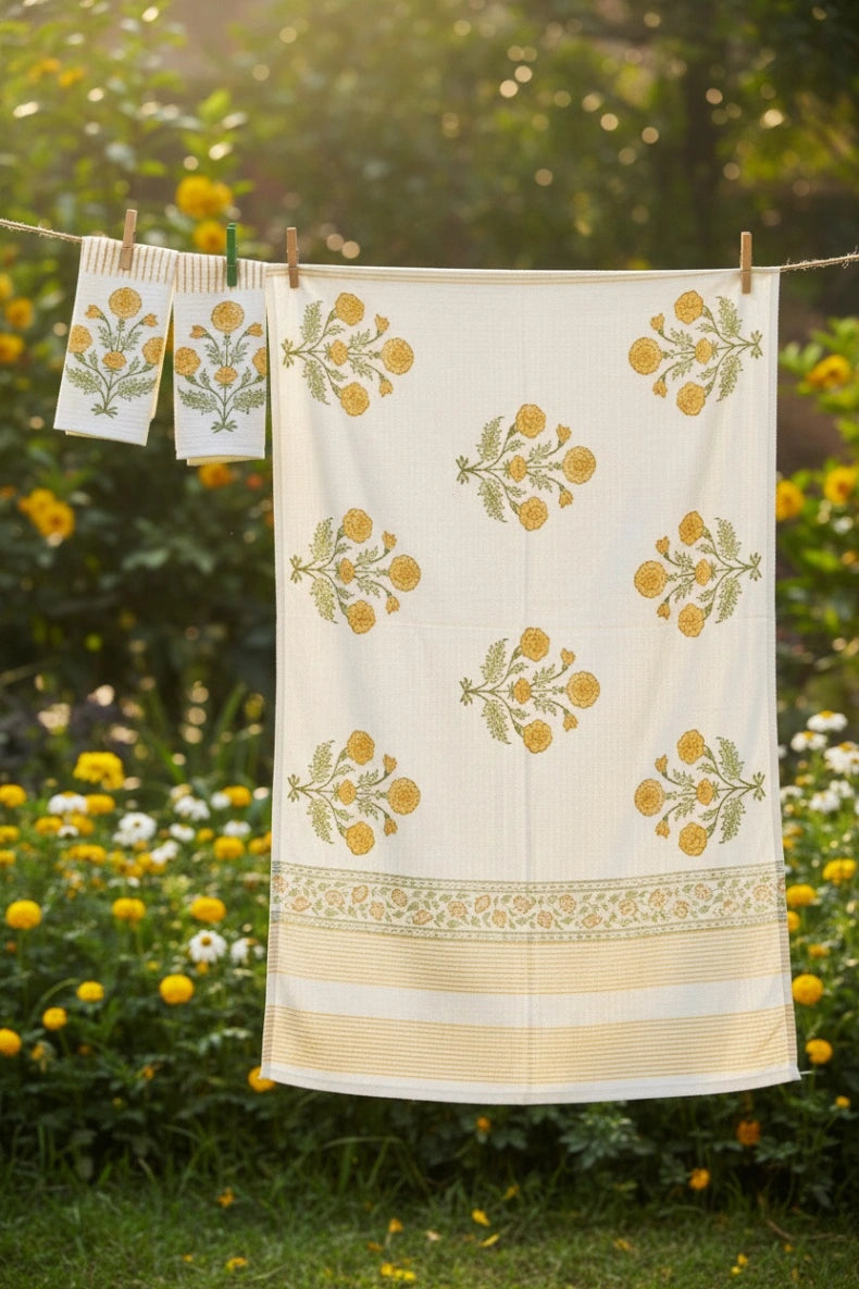 A cream-colored cotton towel with yellow and green floral block prints hangs on a clothesline outdoors in a garden with yellow flowers.