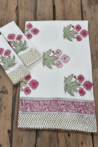 A white cotton textile set, likely a napkin and matching napkin, featuring pink floral prints, green leaves, and decorative borders including chevron and floral patterns, displayed on a rustic wooden background.