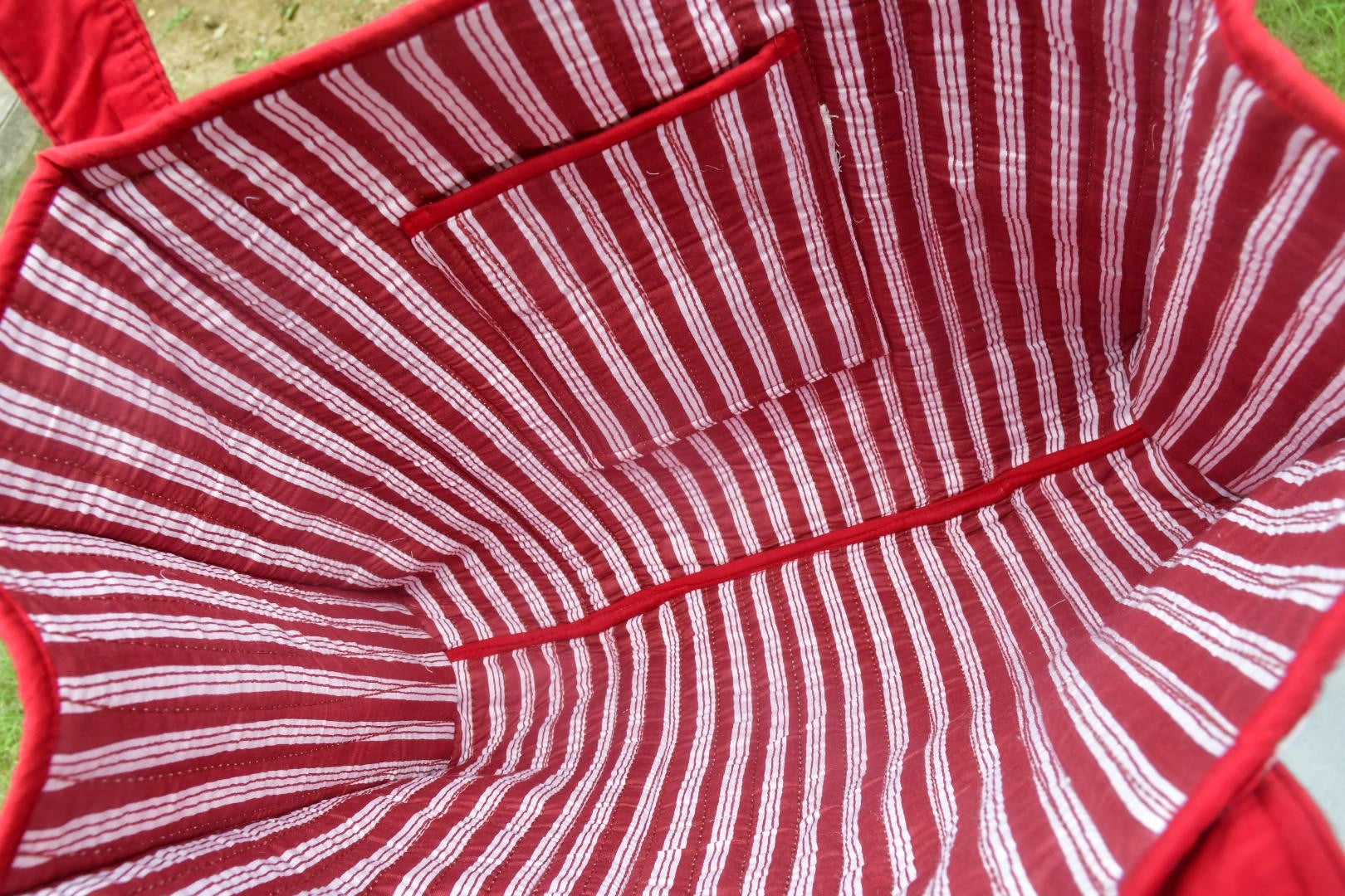 Close-up of the interior of a red and white striped fabric tote bag, showing stitched lining and a top pocket, photographed outdoors on grass.