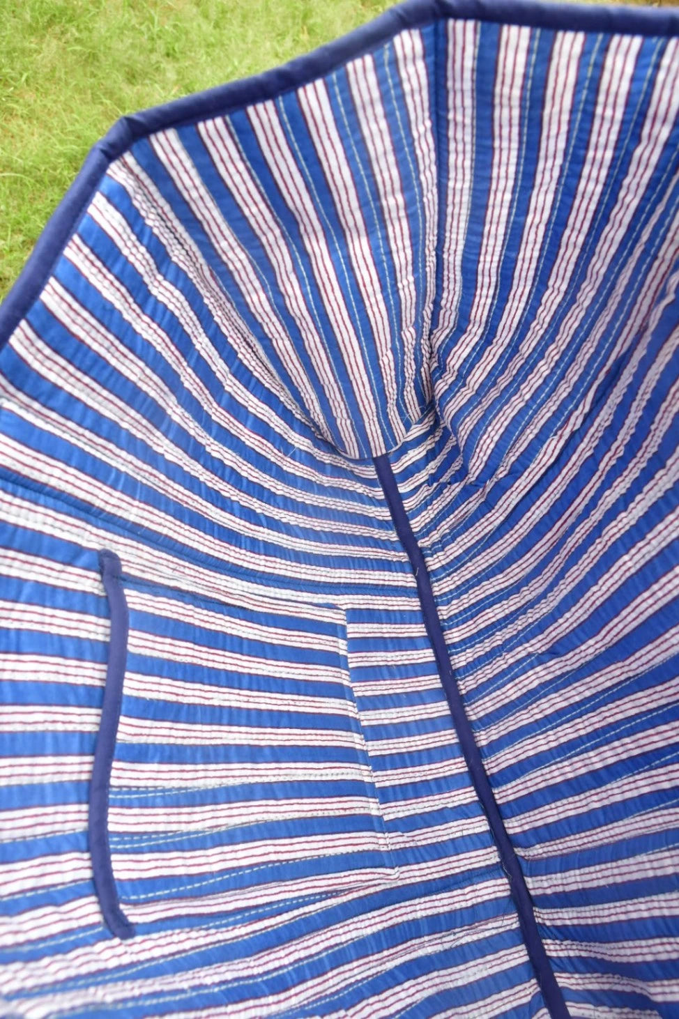 Close-up of the inside of a striped blue and white fabric umbrella, showing the stitched canopy lining and a small pocket, with green grass visible in the background.