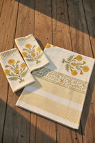 Three cream-colored cotton kitchen towels with yellow and green floral embroidery and striped borders, arranged on a rustic wooden deck with warm sunlight and shadows.