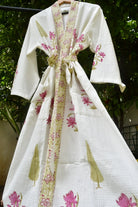 A white waffle-knit kimono robe with pink floral prints and a beige patterned border, hanging on a wooden hanger against a background of green foliage.