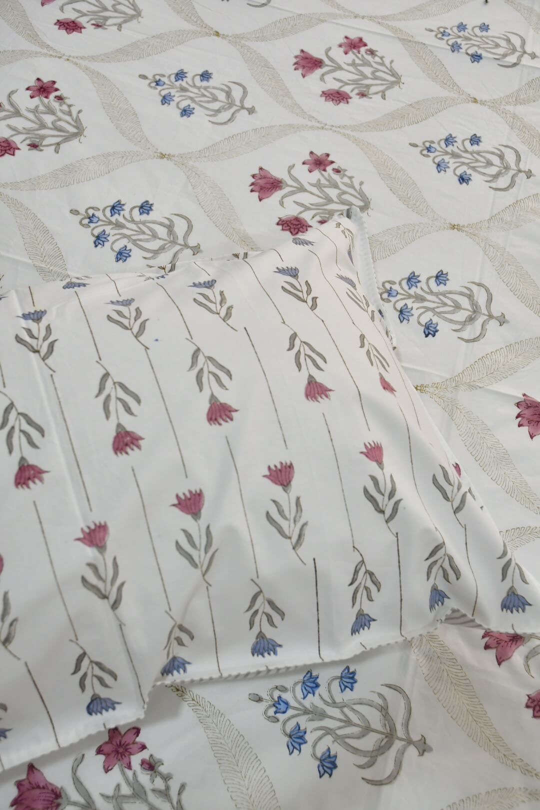 A close-up of a white or off-white fabric textile with a repeating floral block-print pattern in muted pink, blue, and gray-green, with a folded matching pillow or pillowcase placed on top.