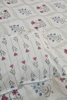 A close-up of a white or off-white fabric textile with a repeating floral block-print pattern in muted pink, blue, and gray-green, with a folded matching pillow or pillowcase placed on top.
