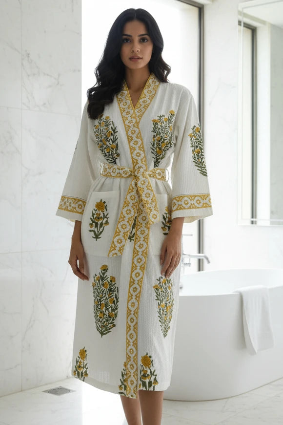 A woman stands in a bright bathroom wearing a white waffle-knit robe with yellow and green floral embroidery and a matching tie belt. A freestanding bathtub and marble walls are visible in the background.