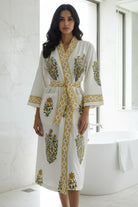 A woman stands in a bright bathroom wearing a white waffle-knit robe with yellow and green floral embroidery and a matching tie belt. A freestanding bathtub and marble walls are visible in the background.