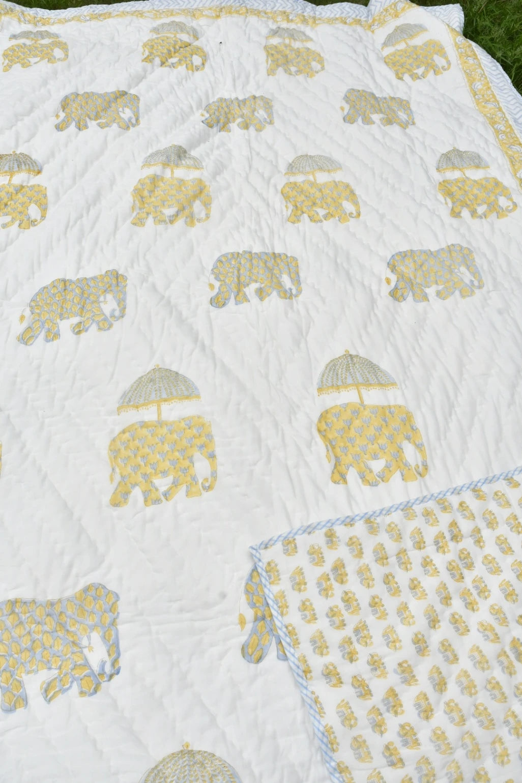 A white quilted blanket featuring a repeating pattern of elephants carrying yellow umbrellas, with a folded corner showing a light blue and white patterned reverse side.