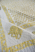 A close-up of a quilted bedcover  featuring a white background with a repeating yellow elephant print, a yellow border with blue paisley motifs, and a large yellow elephant applique on the corner.