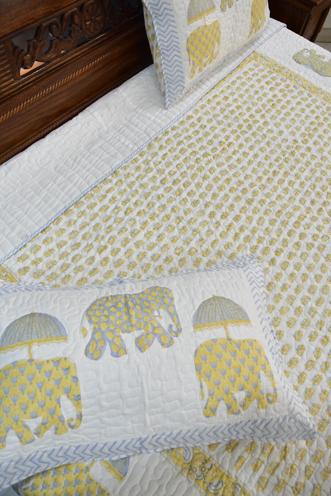 A quilted bedding set featuring a white quilt with a yellow elephant pattern, matching white pillows with blue chevron borders and yellow elephant prints, and a dark wood headboard.