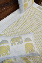A quilted bedding set featuring a white quilt with a yellow elephant pattern, matching white pillows with blue chevron borders and yellow elephant prints, and a dark wood headboard.