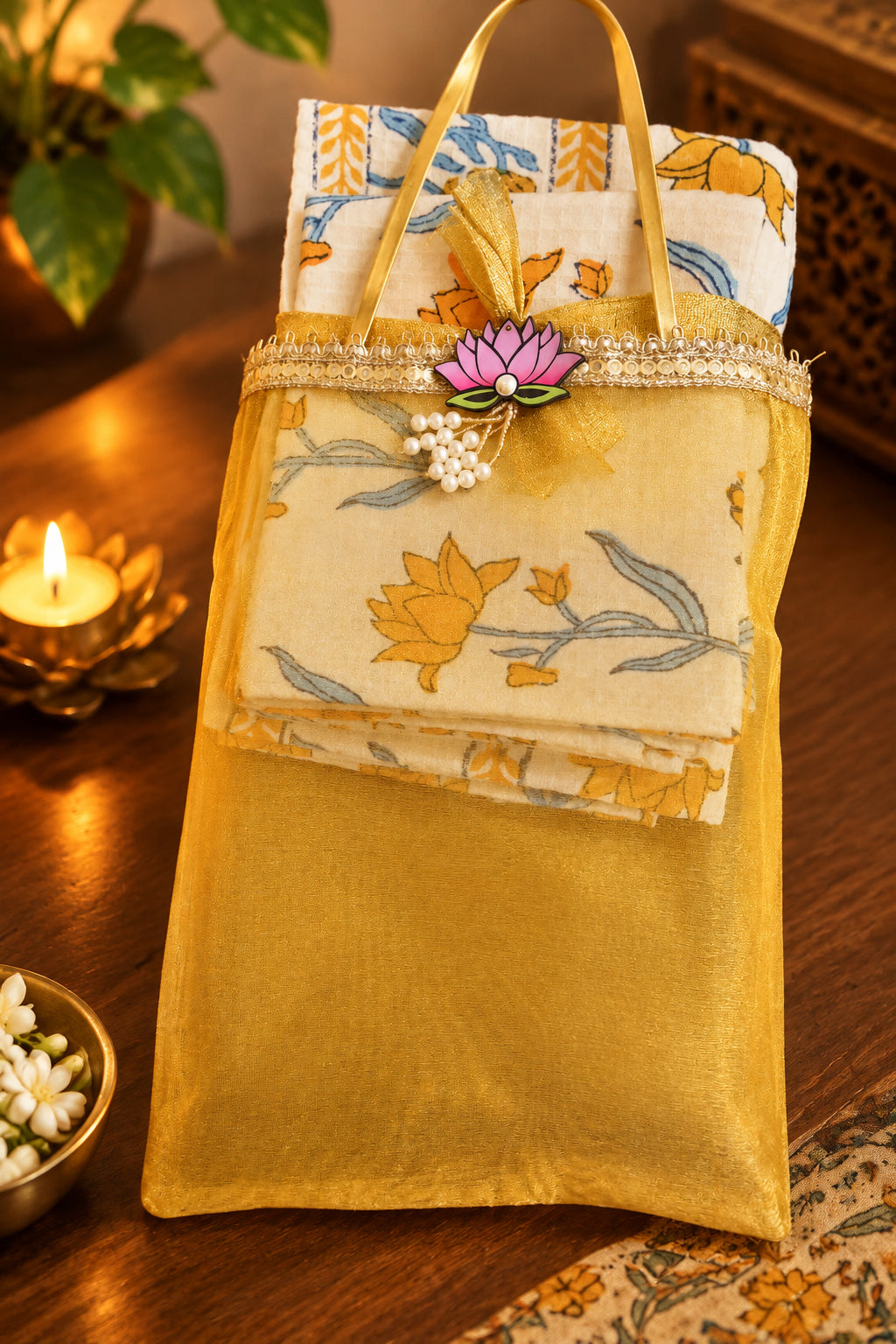 A folded cream-colored fabric cloth with yellow and blue floral patterns, presented inside a yellow mesh carrying pouch with gold handles. The pouch is decorated with a pink lotus flower ornament and a beaded trim, resting on a warm wooden table with a lit candle in the background.