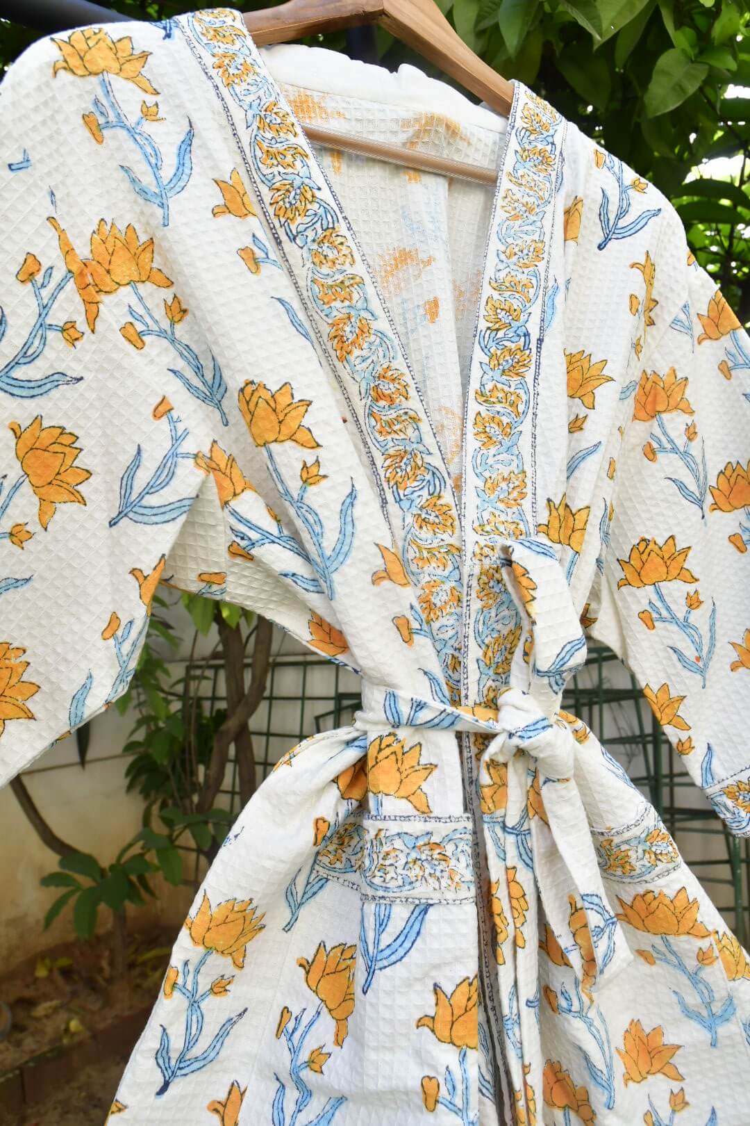 A white cotton kimono robe with a yellow and blue floral print, hanging on a wooden hanger against a background of green foliage.
