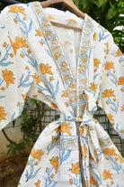 A white cotton kimono robe with a yellow and blue floral print, hanging on a wooden hanger against a background of green foliage.