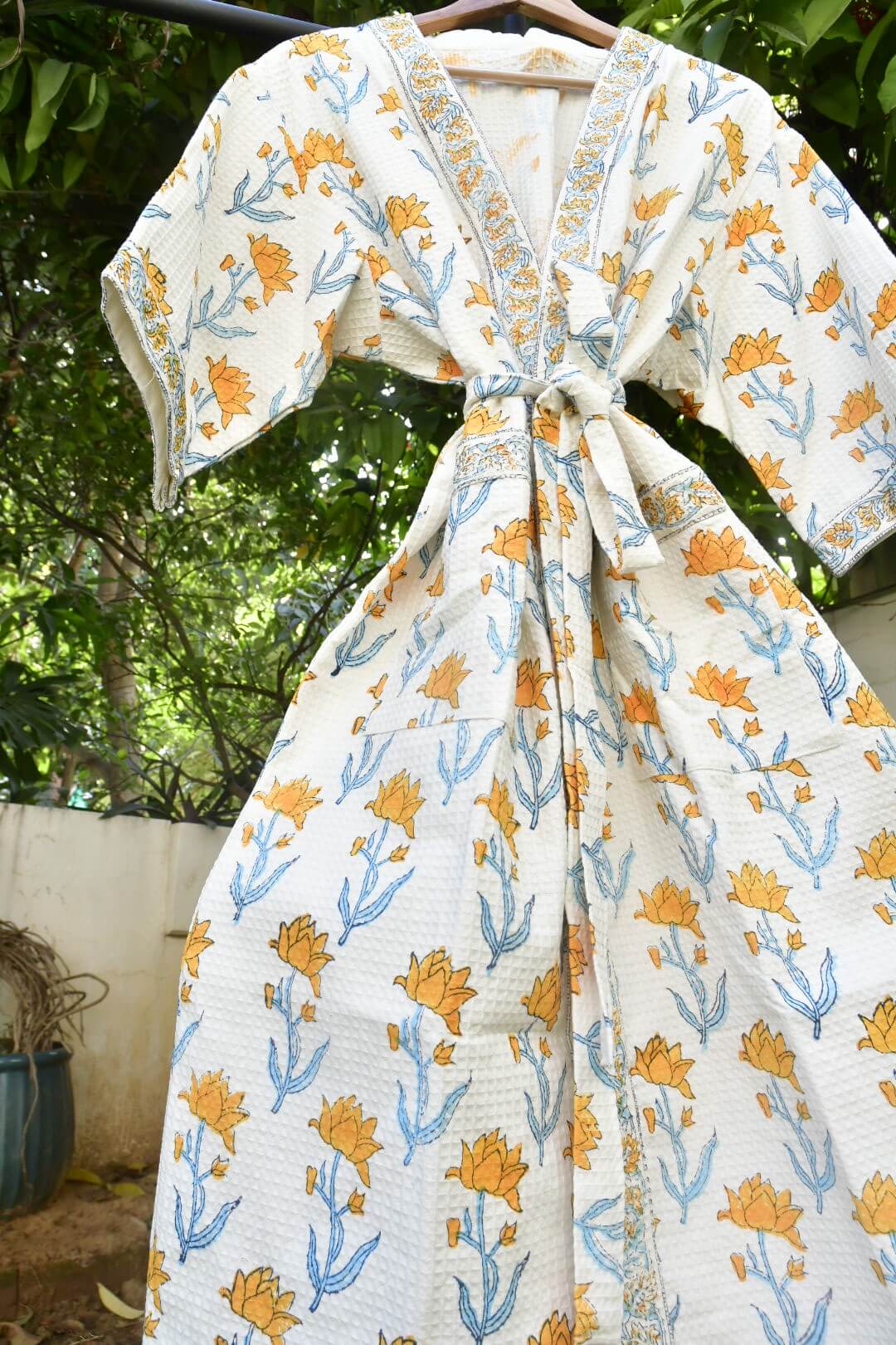 A white cotton kimono robe with a yellow and blue floral block print pattern, hanging on a wooden hanger outdoors against a background of green foliage and a white wall.