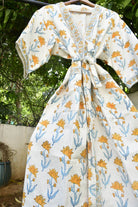 A white cotton kimono robe with a yellow and blue floral block print pattern, hanging on a wooden hanger outdoors against a background of green foliage and a white wall.